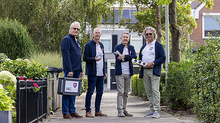 In een straat staan vier mannen op een rij, waarvan de linkerman een koffer draagt. Om de mannen heen ligt een tuin, staat een huis en bevinden zich bomen en struiken.