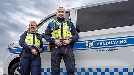 Op de foto zijn twee personen voor een wagen te zien. Het zijn twee handhavers die gele, blauwe en zwarte kleuren dragen. Het gaat om links een vrouw en rechts een man.