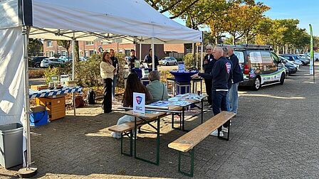 Op de foto staan een kraam met een wit zijl er bovenop en een houten uitklaptafel. Onder de kraam staan mensen.