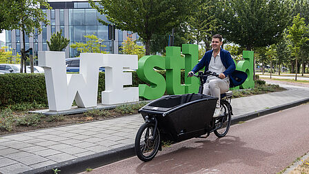Dave van Koppen Westland fiets Op de foto fietst een man met een bakfiets langs grote witte en groene letters met 'Westland' erop