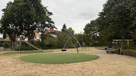 Op de foto is een speeltuin te zien, waarop natuurgras geel is en het kunstgras groen is. Er zijn meerdere speeltoestellen te zien. Er staan ook bomen op de foto.