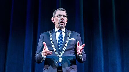 Op de foto spreekt een mannelijke burgemeester met een zwart decor op de achtergrond mensen toe.