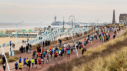 Op de foto zien we een aantal mensen hardlopen aan het strand. Op de achtergrond is een pier te zien. Ook zijn duinen te zien.
