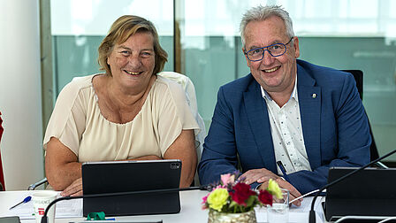 Op de foto zitten een vrouw en een man naast elkaar aan een tafel. Ze kijken recht de camera in.