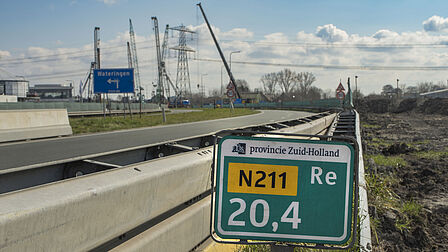Op de foto si een groot groen bood te zien waarop een geel vlak met zwarte letters en verder witte letters staan. Op de achtergrond zie je werkzaamheden aan een weg met allerlei machines om heen.