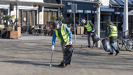 Op de foto rapen mensen met gele hesjes zwerfvuil van de straat op. Dat doen zij met een grijper.