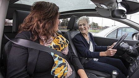 Op de foto ziet u twee vrouwen in een auto zitten, waarbij een senior achter het stuur zit.