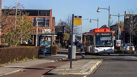 Op de foto rijdt een rood-witte bus door een straat met een kerk en een bushalte op de achtergrond