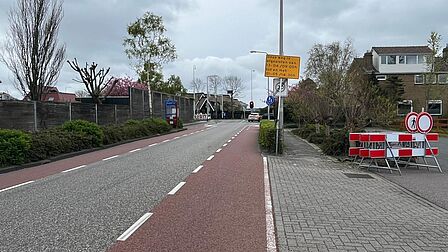 Op de foto zijn een autoweg, twee fietspaden, een lantaarnpaal, een rood-wit hek met bord en een geel bord te zien. Op het gele bord staat dat deze weg een aantal weken is afgezet.