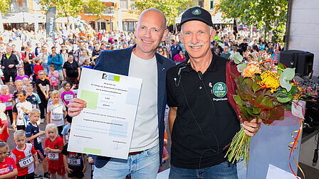 Op de foto staan twee mannen naast elkaar met bloemen, een oorkonde en een mensenmassa op de achtergrond. De foto is vanaf een podium gemaakt.