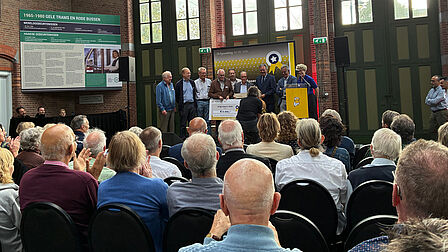 Op de foto is gemaakt in een kerk. Daarop zie je mensen zitten. Zij kijken naar een podium. Ook is links een groen-wit bord te zien.