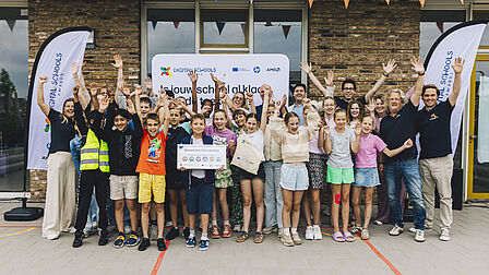Op de foto staan meerdere kinderen en volwassenen op een rij. Zij hebben hun handen in de lucht. Zij staan bovendien voor een schoolgebouw.