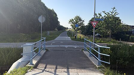 Op de foto zie je twee wegen die elkaar kruisen met een lantaarnpaal en borden. Ook zijn bomen en een brug te zien.