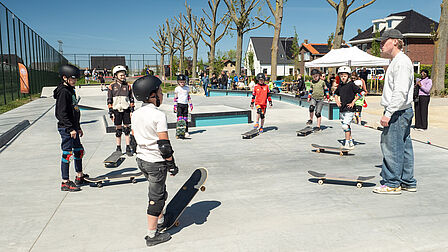 Op de foto staan meerdere kinderen in een kring met beschermende spullen aan en een skateboard aan hun voeten. Een man met een pet en een witte trui geeft hen instructies.