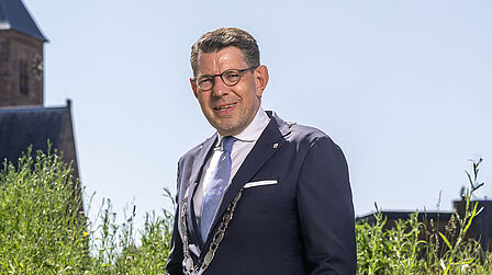 Op de foto staat een man voor planten. Voor hem staan bloemen. Op de achtergrond staat een kerk. De lucht is blauw.