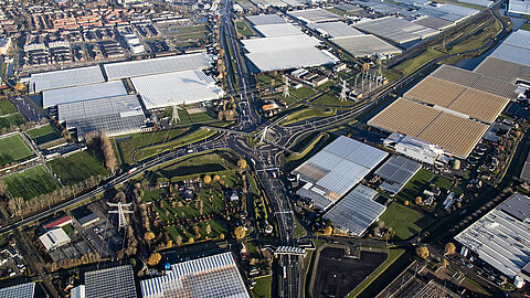 De foto is vanuit de lucht genomen. Daarop zijn kassen, hoogspanningsmasten, wegen, bomen en huizen te zien.