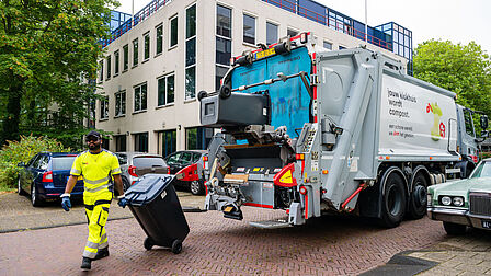 Op de foto zie je een vuilniswagen met daarvoor een man die een minicontainer in zijn handen heeft.