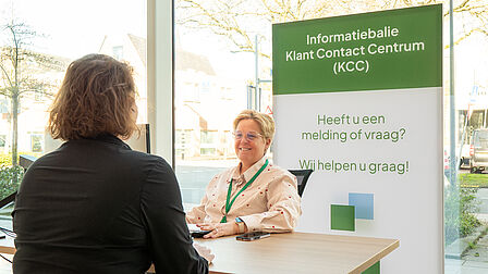 Op de foto helpt een vrouw aan een tafel een andere vrouw. Verder is een wit-groen bord met tekst erop te zien.