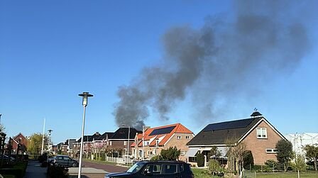 Op de foto is een zwarte rookpluim te zien, met huizen, een straat, bosjes en een lantaarn paal op de voorgrond. De lucht is blauw.