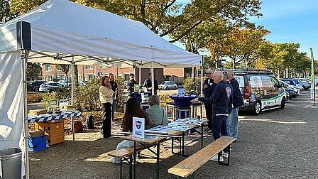 Op de foto staan meerdere mensen onder een tent en zijn meerdere bomen te zien.