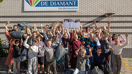 Op de foto staan meerdere kinderen bij elkaar die een vuilniszak in de lucht houden of handen in de lucht steken. Tussen de kinderen staan meerdere volwassenen die meejuichen.