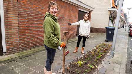 Twee vrouwen poseren op een foto met tuingereedschap in hun hand en meerdere bloemen in een plantvak.